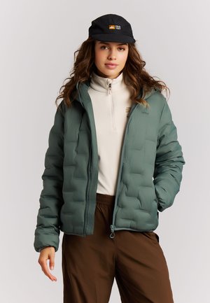 Jonge vrouw met golvend haar, een zwarte pet, groene gewatteerde jas, beige top met rits en een bruine broek tegen een effen achtergrond.