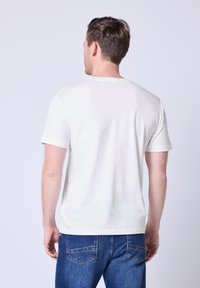 Biała bawełniana koszulka z okrągłym dekoltem i krótkimi rękawami, zestawiona z niebieskimi dżinsami. Widoczne przeszycia i detal szwu na tylnej części ramienia.