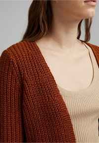 Rust knit cardigan in a warm reddish-brown color, featuring a relaxed fit and open front. Worn over a light beige ribbed tank top.