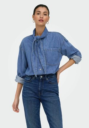 Kvinde iført langærmet denimskjorte med opkrogede ærmer og slips, kombineret med højtaljede mørkeblå jeans, stående med den ene hånd på hoften.