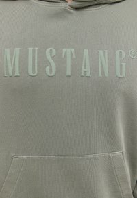 Sweat à capuche vert avec une poche avant, arborant un texte en relief "MUSTANG" dans une teinte assortie. Tissu doux et texturé avec une coupe décontractée.