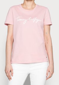Světle růžové bavlněné tričko s krátkým rukávem, které má bílý kurzívní nápis "Tommy Hilfiger" přes hruď. Kulatý výstřih.