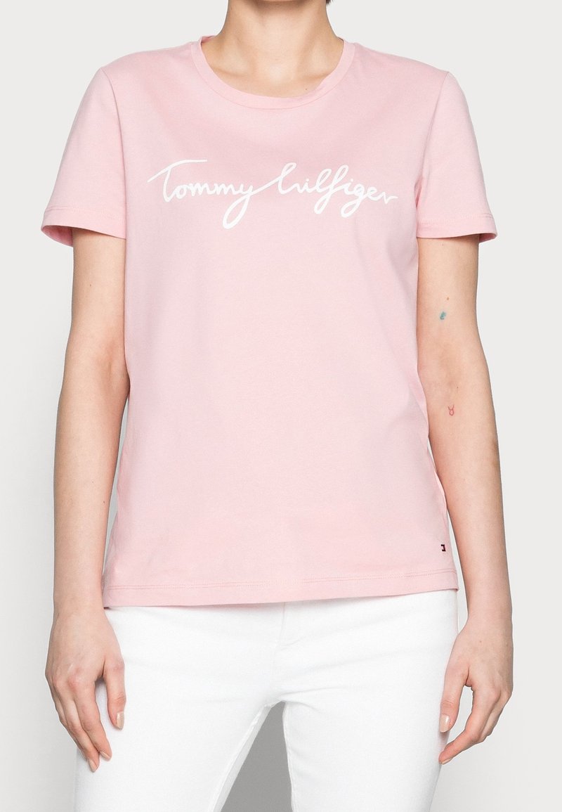 Světle růžové bavlněné tričko s krátkým rukávem, které má bílý kurzívní nápis "Tommy Hilfiger" přes hruď. Kulatý výstřih.