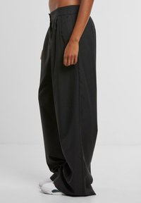 Personne portant un pantalon noir large taille haute et des baskets blanches, debout devant un fond uni clair.