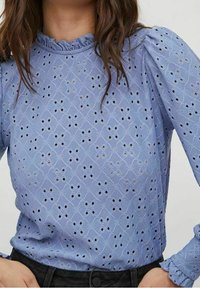 Vaaleansininen paita, joka on valmistettu silkkikankaisesta kankaasta, jossa on rypytetty kaula-aukko ja puffattuja hihoja. Suunnittelussa on monimutkainen kuvio, jossa on pieniä lävistyksiä.