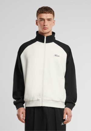 Men's zip-up jacket in white and black, featuring a high collar, elastic cuffs, and two side pockets. Soft texture with logo detail on chest.