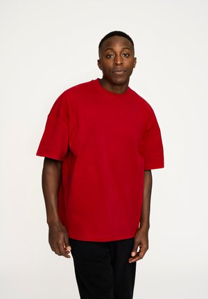 Rød oversized t-shirt lavet af blødt stof, med rund halsudskæring og korte ærmer. Båret med sorte bukser, sat mod en ensfarvet baggrund.