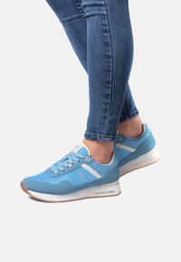 Vaaleansiniset lenkkarit, joissa on yhdistelmä sileitä ja teksturoituja materiaaleja, valkoiset yksityiskohdat, nauhallinen malli ja kumipohja.