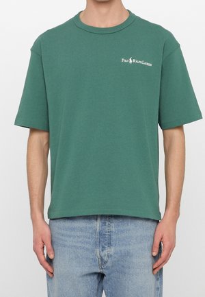 Mężczyzna ubrany w zielony t-shirt Polo Ralph Lauren z krótkimi rękawami i jasnoniebieskie dżinsy, stojący na jednolitym tle.