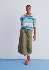 Pullover de fleece azul com riscas laranja, combinado com uma saia longa verde-oliva. O modelo usa meias brancas e ténis castanhos.
