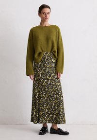 Vrouw in een olijfgroene gebreide trui en een gestreepte lange rok in zwart, wit en mosterd met zwarte instappers, staande tegen een witte muur.