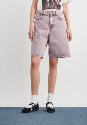 Személy világoslila farmer térd alatti rövidnadrágban, fehér zokniban, fekete balettcipőben és fehér, grafikás pólóban áll kék szőnyegen.