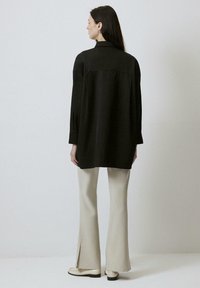 Black long-sleeve shirt with a collar, loose fit, back yoke detail; paired with cream flared trousers and white shoes, minimalistic design.