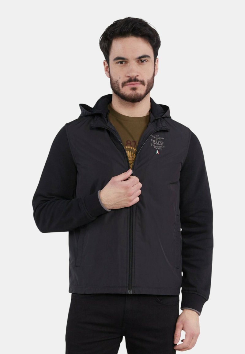 Giacca nera con zip e cappuccio, caratterizzata da una combinazione di texture lisce e a coste, un logo discreto sul petto e tasche laterali.