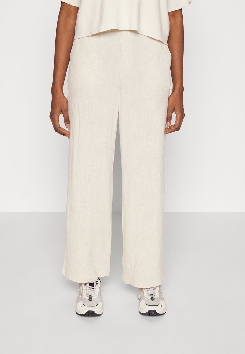 Pantalones de lino anchos color beige con un acabado texturizado, que presentan un corte relajado y se combinan con zapatillas ligeras.