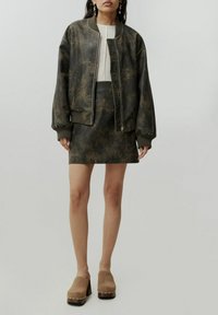 Olivengrüne Bomberjacke mit verwitterter Textur, gerippten Bündchen und Saum, kombiniert mit einem passenden Leder-Minirock und beigen Plateausandalen.