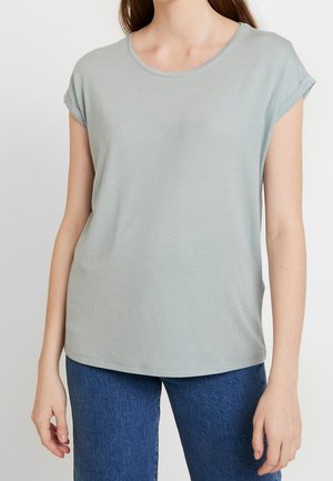 Kvinna i en ljusgrå kortärmad t-shirt med uppvikt ärm och blå jeans, som står mot en helt vit bakgrund.