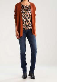 Brązowy kardigan w prążki na czarnej bluzce w kwiaty, zestawiony z ciemnoniebieskimi dżinsami i czarnymi botkami za kostkę. Neutralne tło.