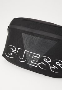Fekete szövet táska sima tapintással, cipzáros záródással, és feltűnő fehér "GUESS" felirattal, szürke háromszög akcentussal.
