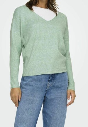 Vrouw die een lichtgroene V-hals trui draagt over een wit shirt, gecombineerd met losse blauwe spijkerbroek, staand tegen een effen achtergrond.