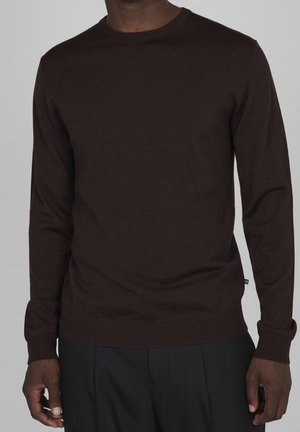 Man wearing a fitted dark brown long-sleeve sweater and black pants, standing against a plain gray background.