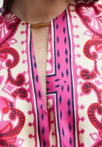 Blouse à motifs avec un design rose et crème, rehaussé de détails bleu foncé, un décolleté en V et un détail en métal doré au niveau du cou.