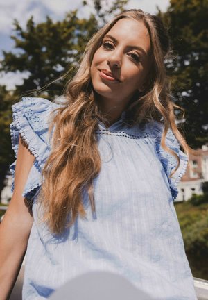 Lichtblauw topje met gerimpelde mouwen, kantafwerking en een verticale streepjespatroon. Model heeft lang, golvend haar en een zachte uitdrukking.