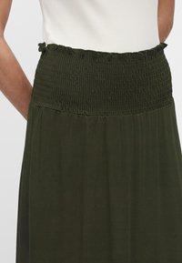 Jupe verte foncée taille haute avec ceinture élastique froncée portée avec un haut blanc sans manches.