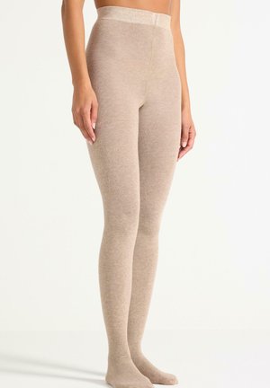 Kvinde iført højtaljede, beige, ribstrikkede leggings, stående på en lys baggrund med hænder afslappet langs siden.