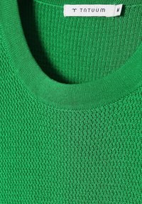 Maglione verde a collo tondo in maglia che mostra un tessuto strutturato e un'etichetta bianca con il nome del marchio e la taglia M.