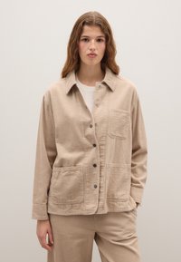 Beige corduroyjas met een knoopsluiting, twee grote voorkruiken en een kraag. Geweven stof met zichtbare ribbels.