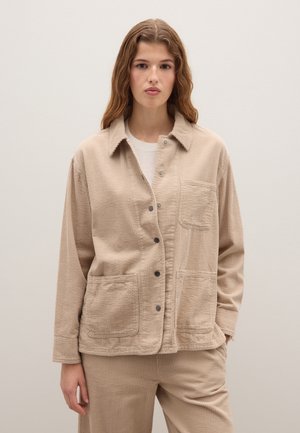 Beige corduroyjas met een knoopsluiting, twee grote voorkruiken en een kraag. Geweven stof met zichtbare ribbels.