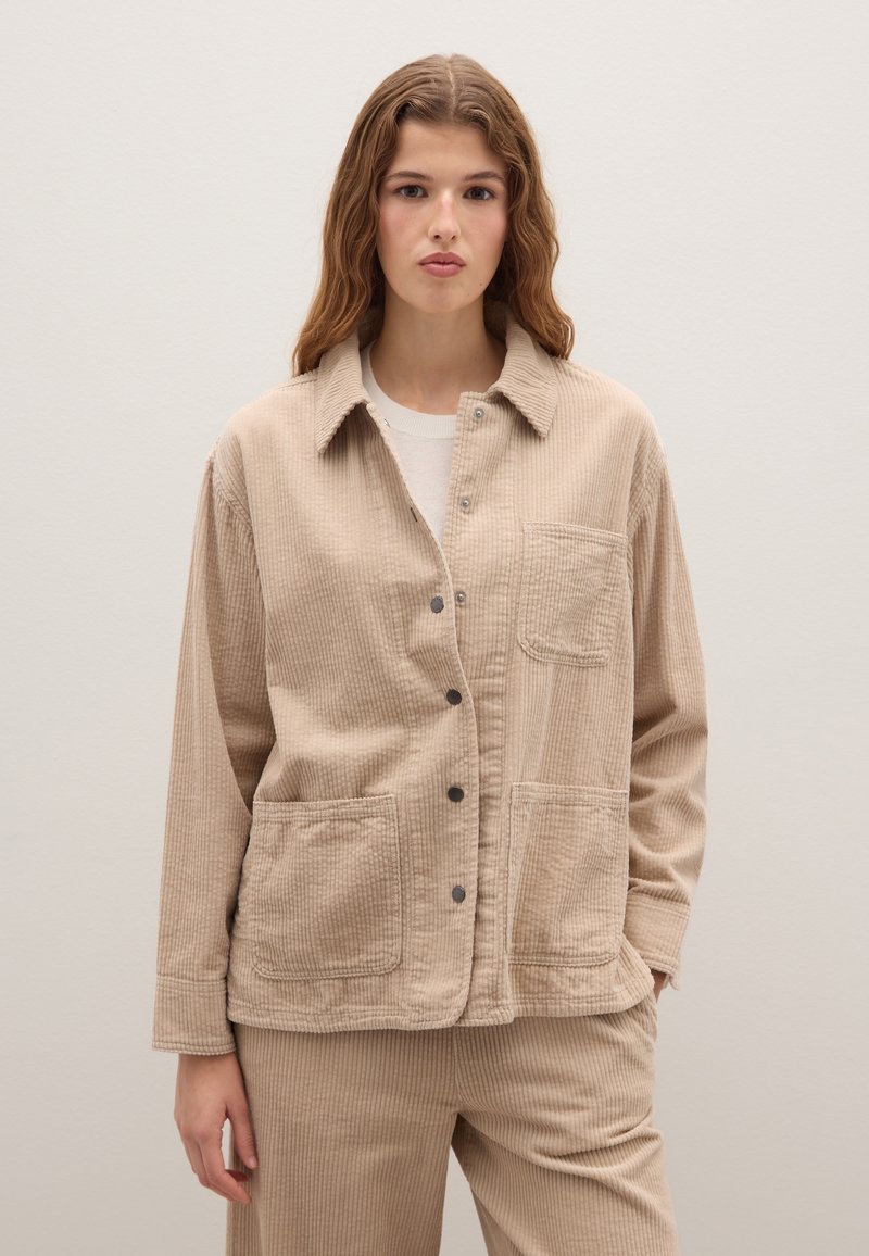 Beige corduroyjas met een knoopsluiting, twee grote voorkruiken en een kraag. Geweven stof met zichtbare ribbels.