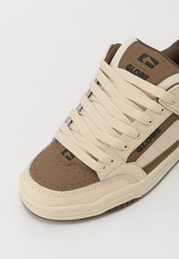 Beige and brown sneaker featuring a thick sole, lace-up design, and logo. Made from synthetic materials with a smooth and textured finish.