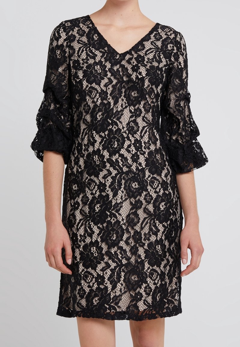 Robe en dentelle florale noire avec superposition de tissu transparent, décolleté en V et manches ballon trois-quarts sur fond neutre.