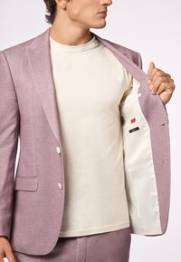 Hellblauer Blazer mit zwei Knöpfen und Reverskragen, kombiniert mit einem cremefarbenen T-Shirt. Innenfutter aus weißem Satin mit Organisationsfächern.