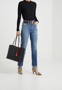 Svart läder tote-väska med röda detaljer, guldkam hardware och slät textur. Modellen bär en ribbad svart topp, blå jeans och ankelboots med ormskinnsmönster.