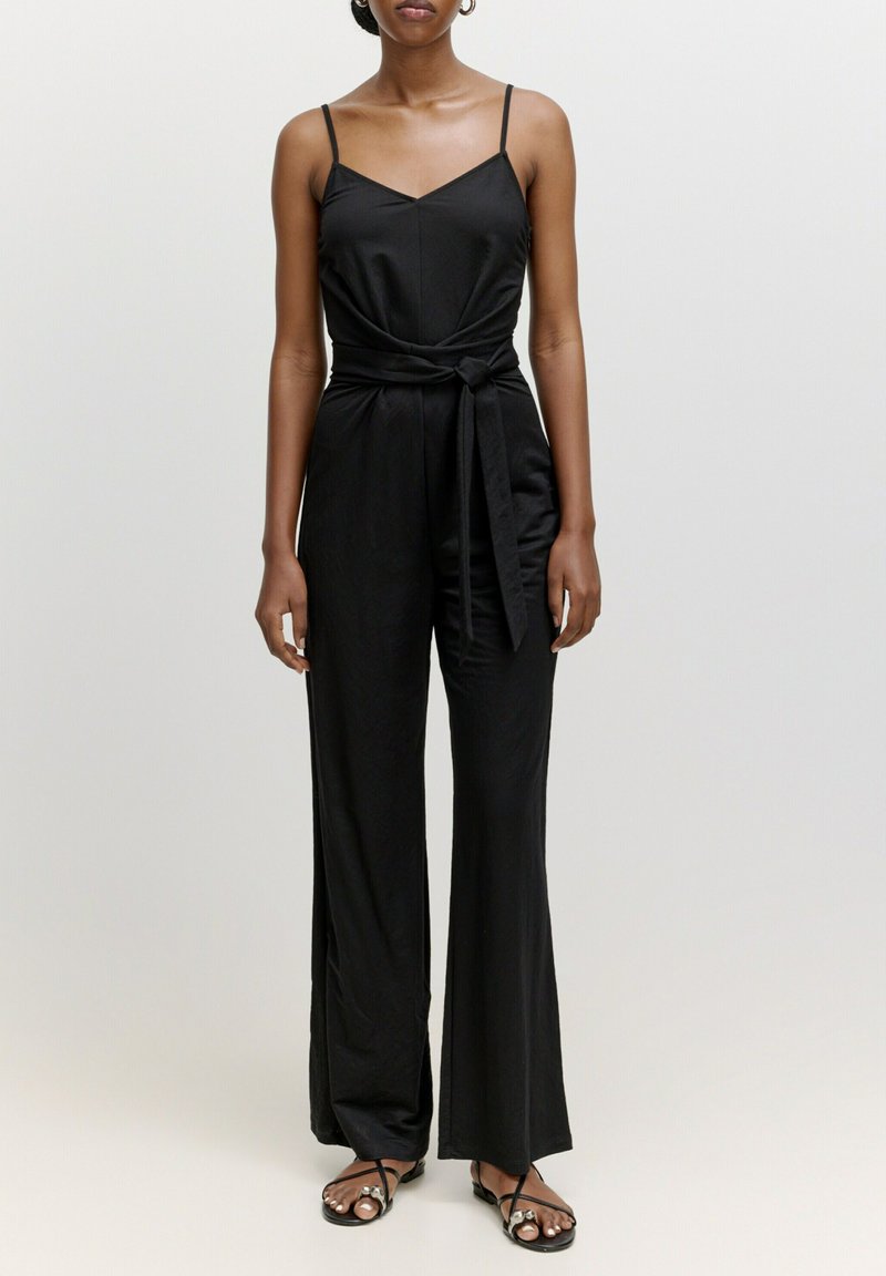 Schwarzer Jumpsuit mit dünnen Trägern, V-Ausschnitt, gebundenem Taillendetail und weit geschnittenen Hosenbeinen. Der Stoff wirkt glatt und hat einen leichten Glanz.