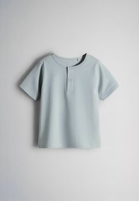 Chemise bleu clair à manches courtes avec col rond et deux boutons sur la patte de boutonnage avant, étalée à plat sur un fond blanc.