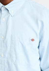 Lyseblå skjorte med knapper og brystlomme, der har et lille Dickies mærkelogo på lommen.