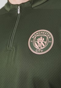 Zöld sportfelső, negyedcipzárral, textúrált anyagból készült. A mellkason rózsaszín Manchester City logó található, sötétzöld háttérrel.