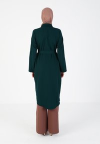 Green, long jacket with a belted waist and long sleeves. Worn over brown wide-leg pants. Background is plain white.
