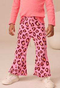 Pantalones acampanados de tela acanalada rosa con un audaz estampado de leopardo en marrón oscuro y naranja. Rematados con un dobladillo ondulado. Combinados con zapatillas blancas.