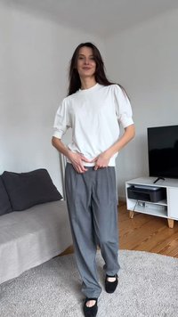 Kvinne står innendørs iført en hvit topp med pufferm, grå løse bukser og svarte sko, smilende med hendene nær livet.