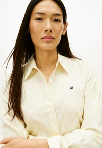 Lichtgeel overhemd met knopen en verticale witte strepen. Heeft een klassieke kraag en een klein geborduurd logo op de linkerborst.