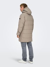 Beige puffer jacket with a hood, featuring a quilted design and a black accent on the sleeve, paired with light blue jeans and white sneakers.