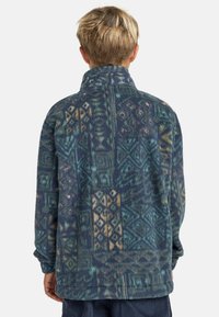 Fleecejacke in blau-grünen Tönen mit geometrischen Mustern, hohem Kragen und langen Ärmeln. Aus weichem, strukturiertem Material.