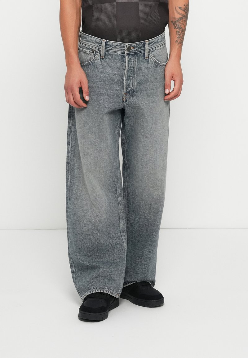 Szerokie, szare dżinsy z wyblakłym wykończeniem, pięć kieszeni, widoczne szwy. Noszone z czarnymi butami na białym tle.