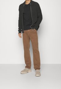 Svart zip-up cardigan, texturerad tyg. Svart t-shirt under. Bruna raka byxor. Beiga skor med rund tå och ljus sula.