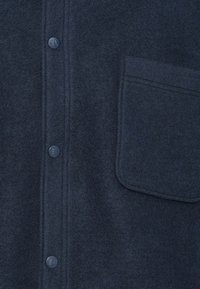 Σκούρο μπλε πουκάμισο από μάλλινο ύφασμα με τρία στρογγυλά κουμπιά και μία μεγάλη τσέπη στο δεξί μέρος.